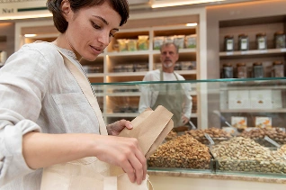 Een vrouw in een licht shirt en met een draagtas kiest producten uit een notenrek in een winkel. Een winkelier in een schort staat achter de toonbank, met planken vol potten op de achtergrond.
